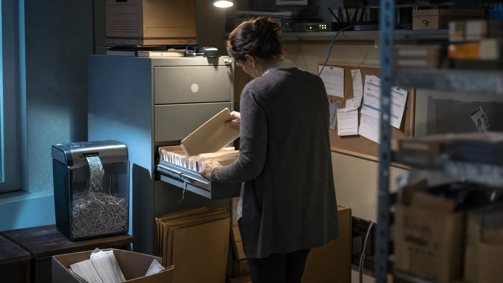 Une femme de dos à la cinquantaine dans un coin archives de boutique en ligne, un tiroir de classeur métallique ouvert, une main soulevant un dossier de formulaires papier, un petit destructeur de documents en marche à côté d'elle sous une ampoule nue, un routeur allumé sur une étagère au-dessus.